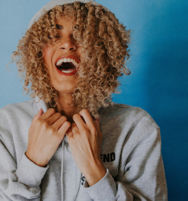 A person with curly hair cascading over their face and a gray hoodie is laughing against a vibrant blue background.