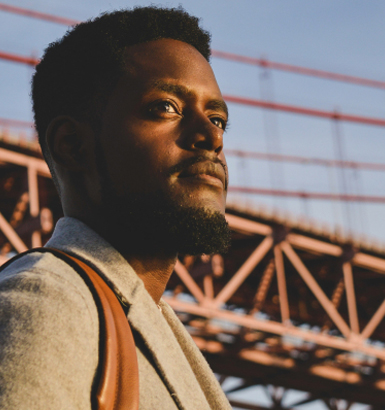 At sunset, a bearded individual stands gracefully before a bridge, wrapped in a gray coat and shouldering a brown bag.