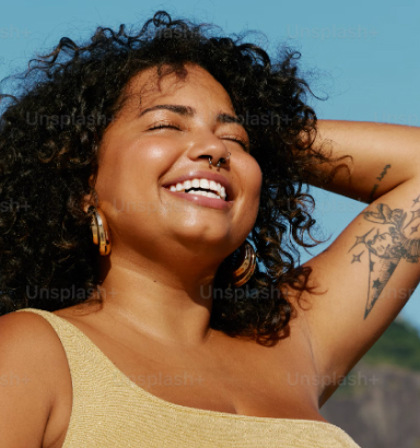 A smiling woman with curly hair and tattoos enjoys the sunlight, wearing a beige sleeveless top amidst a serene outdoor setting.