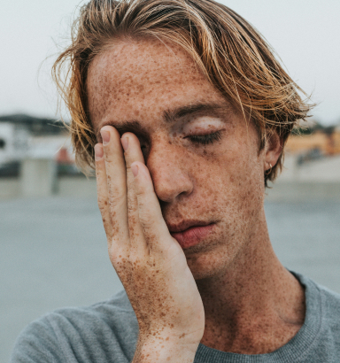 A freckled individual stands outdoors, one hand shielding their weary face from the elements.