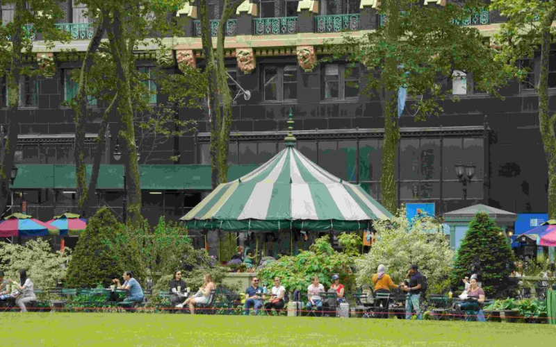 People sitting on benches near a green-striped tent in a park with trees and a building in the background.