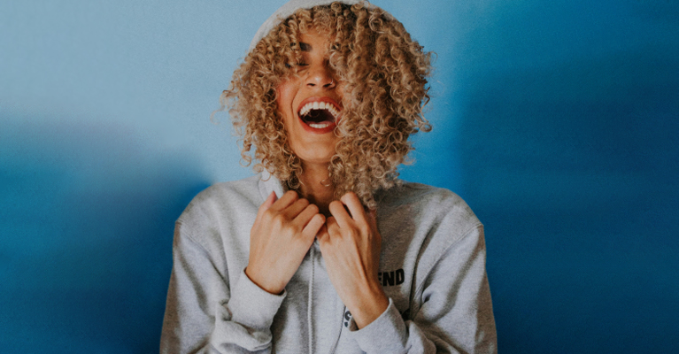 Person with curly hair laughing, wearing a gray hoodie against a blue background.