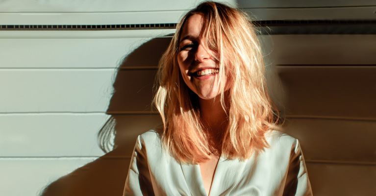 Woman with blonde hair smiling in sunlight against a light-colored wall.