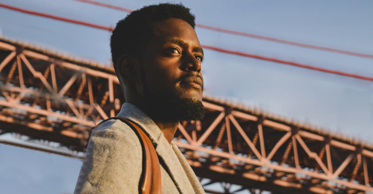 A person in a gray coat stands outdoors with a red bridge in the background, looking into the distance.
