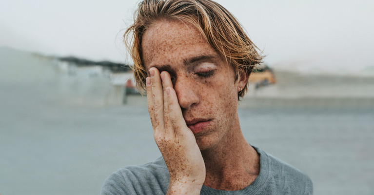 A young person with freckles rubs their eye, appearing tired, against an outdoor blurry background.