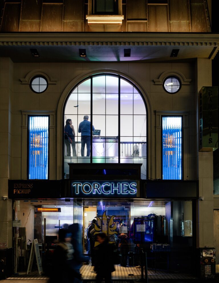 City building at night with large arched window, two people inside, and a neon sign reading Torches above the entrance.