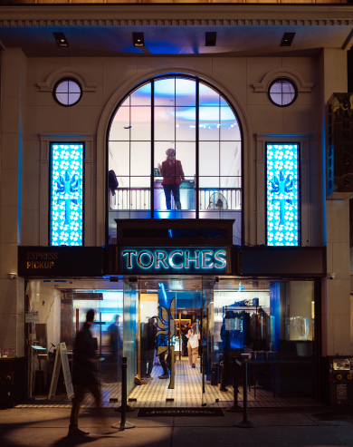 Night view of a restaurant entrance with a neon TORCHES sign and a person standing on the upper level inside.
