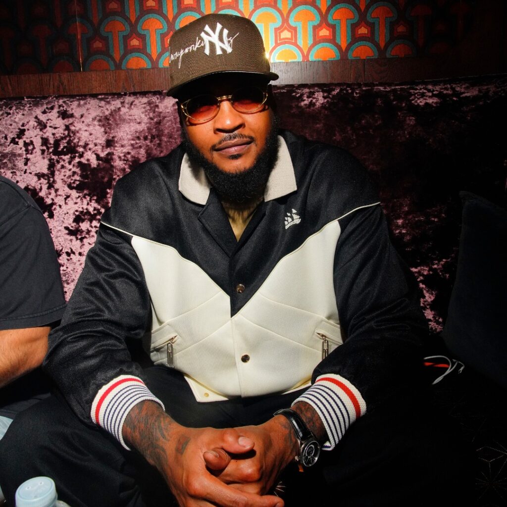 Man wearing a brown New York Yankees cap, STAYME7O sunglasses, and a black-and-white jacket sits on a velvet couch.