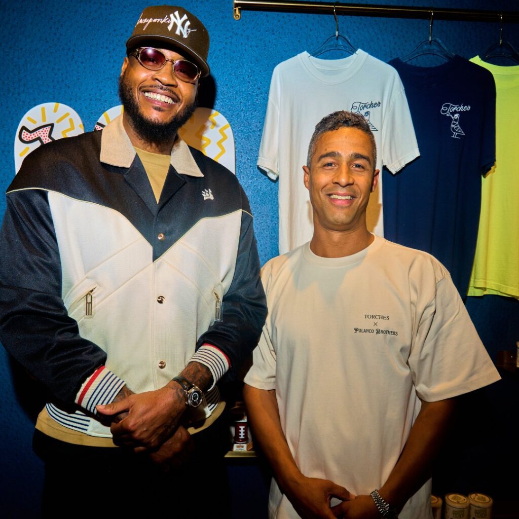 Two men smiling and posing together in front of a display of STAYME7O T-shirts on a blue wall.
