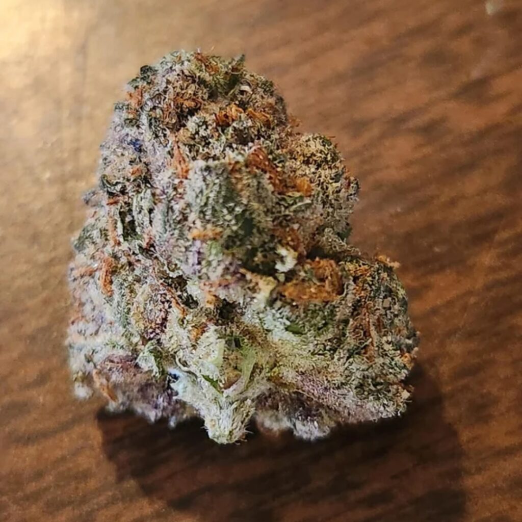 A close-up of a cannabis bud with orange hairs and frosty trichomes on a wooden surface.