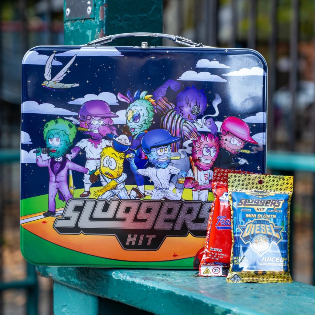 A colorful lunchbox with cartoon characters and two snack bags labeled Sluggers HIT on a table outdoors.