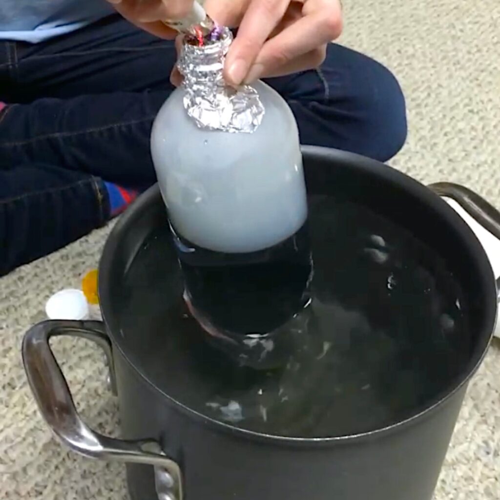 A hand holds a foil-covered bottle with smoke over a pot of water on a carpeted floor.
