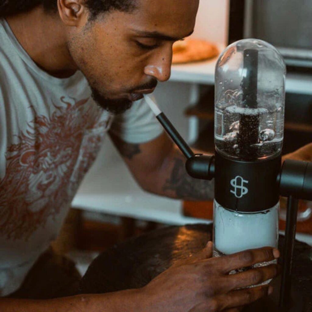 A person uses a glass water pipe, inhaling through a mouthpiece while smoke fills the chamber.